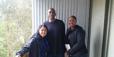 Three people standing on a balcony with a corrugated metal wall and trees in the background.